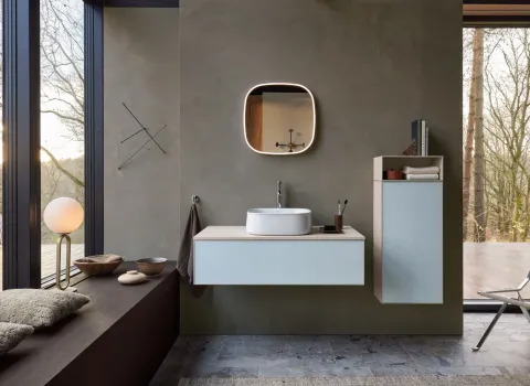 Ein modernes Badezimmer mit minimalistischer Einrichtung und großem Fenster bietet einen Blick in den Wald. Die helle, schwebende Waschtischkonsole mit einem weißen Aufsatzwaschbecken steht im Mittelpunkt, darüber hängt ein beleuchteter, quadratischer Spiegel. Rechts davon ist ein schmaler Hochschrank mit Handtüchern und Dekorationen montiert. Die natürliche Farbgestaltung unterstreicht die ruhige Wellness-Atmosphäre. Fokus-Keyword: modernes badezimmer